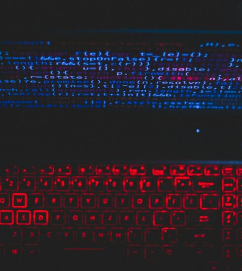 Dark-themed laptop setup with a red glowing keyboard and code on screen, ideal for tech enthusiasts.